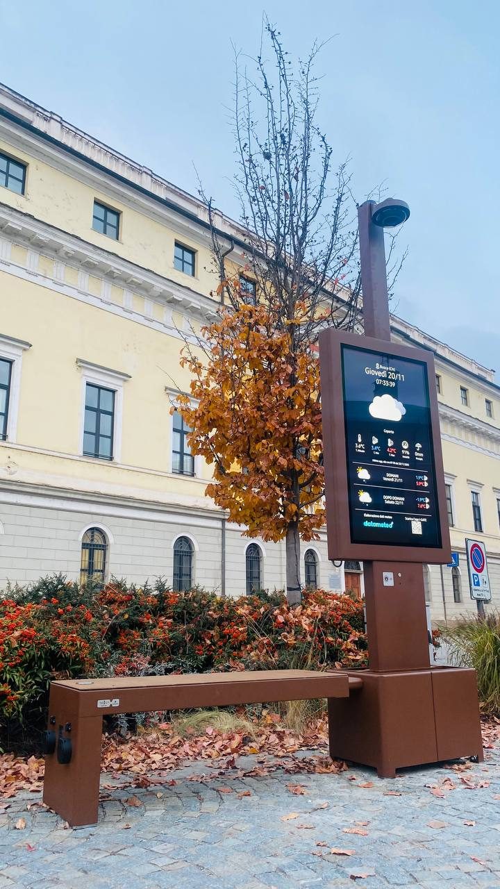 Isola digitale i-Gate con display e panchina smart davanti a un palazzo storico in autunno
